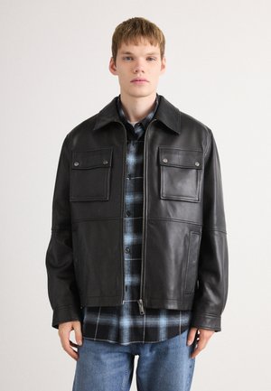 Black leather jacket with a collared neckline, front zipper, two chest pockets, and a loose fit, layered over a plaid shirt and denim jeans.