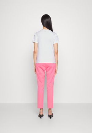 Woman standing facing away wearing a white short-sleeve shirt, pink pants, and black heeled shoes against a plain light background.