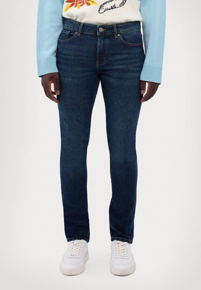 Dark blue denim jeans with a slim fit, featuring five pockets, a zip fly, and contrasting stitching details, paired with white sneakers.