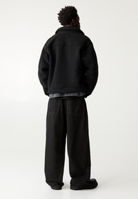 Veste en polaire noire surdimensionnée avec un col montant, portée sur une chemise grise. Associée à un pantalon en denim noir large et des chaussures noires.
