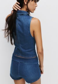 Denim sleeveless top in dark blue with a form-fitting design, featuring a high collar and visible seams. Paired with matching shorts.