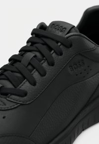 Baskets en cuir noir avec lacets noirs, embossées du logo "BOSS" sur le côté et la languette, présentant un design de panneaux texturés et lisses.