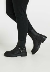 Black ankle boots with a matte finish, featuring a side zipper, chunky rubber sole, and a buckle strap detail.