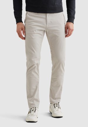 Moški, ki nosi svetlo bež ozke hlače, bele visoke superge in črno dolg rokav, stoji na enobarvni podlagi.