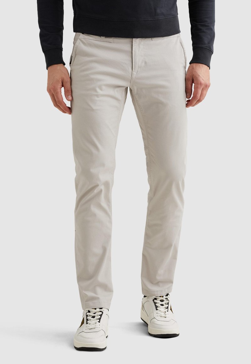 Mann trägt hellbeige Slim-Fit-Hose, weiße High-Top-Sneaker und ein schwarzes langärmeliges Oberteil, steht vor einem einfarbigen Hintergrund.