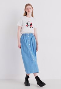 Maglietta bianca con design grafico di due personaggi, abbinata a una gonna maxi in denim blu chiaro con bottoni frontali e vita elastica.