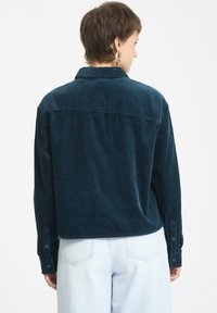 Tealfärgad manchester skjorta med vertikala ränder, knäppning vid ärmsluten och en skräddarsydd ryggdesign. Bärs med ljus denim.