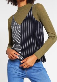 Flerfärgad topp med en mörkgrön långärmad tröja under en ärmlös överdel i marinblått och vitt randigt mönster, med volanger.