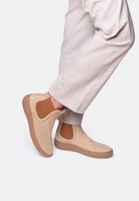 Stivali alla caviglia beige in suede con pannelli laterali elastici a coste, abbinati a pantaloni beige chiaro, che mostrano un design pulito e semplice.