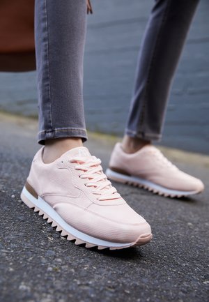 Persoon met lichtroze sneakers en grijze skinny jeans loopt op een gestructureerd asfaltoppervlak buiten.
