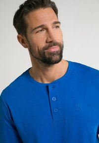 Modrá košile s dlouhými rukávy a kulatým výstřihem, s placketem na tři knoflíky a embosovaným logem. Měkká bavlněná tkanina s hladkým povrchem.