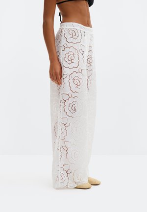 Külgvaade inimesest, kes kannab läbipaistvaid valgeid lillekübaraid pükse, musta bikiini ülaosa ja beežid punutud teravnäolisi kingi tavalise tausta ees.