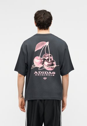 Man draagt een donkergrijs Adidas Originals t-shirt met een roze afbeelding van drie kersen en de tekst "Sweet Cherries" op de achterkant.
