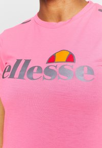 Camiseta de algodón rosa con un gran logotipo gris "ellesse" y un acento en naranja y amarillo encima. Mangas cortas, cuello redondo, textura suave.