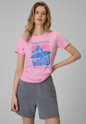 Kobieta nosząca różowy t-shirt z niebieskim nadrukiem surfingowym i napisem, w połączeniu z szarymi casualowymi szortami, stojąca na jednolitym tle.