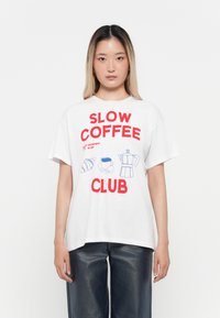 Jauna sieviete, kas valkā balto T-kreklu ar sarkanu tekstu "Slow Coffee Club" un zilu ilustrāciju par kruasānu, kafijas krūzi un kafijas automātu.
