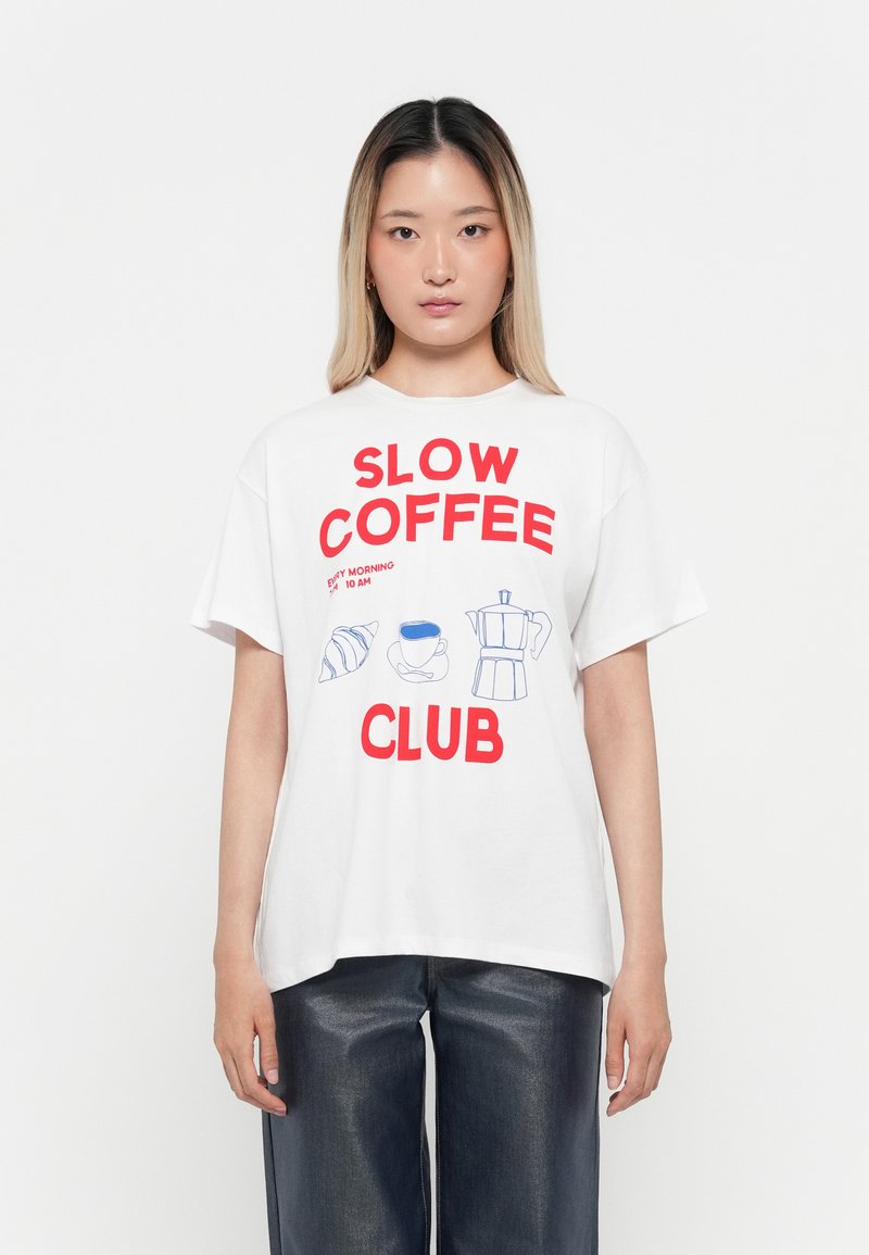 Jauna sieviete, kas valkā balto T-kreklu ar sarkanu tekstu "Slow Coffee Club" un zilu ilustrāciju par kruasānu, kafijas krūzi un kafijas automātu.