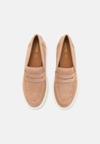 Chaussures en daim à enfiler de couleur rose clair avec un bout cousu, un détail décoratif à l'avant et une semelle blanche contrastante. Nom de la marque visible à l'intérieur.