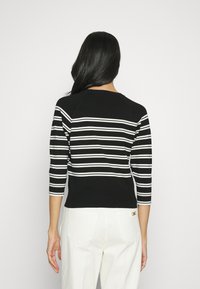 Kobieta odwrócona tyłem, ubrana w czarno-białą, paskowaną bluzkę z długim rękawem i białe spodnie, na tle jasnego, jednolitego tła.