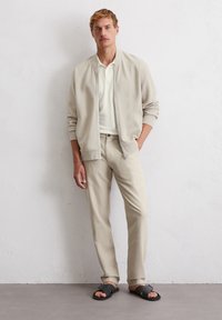 Light beige bomber jacket over a white polo shirt, paired with light beige trousers and black slide sandals, against a neutral background.
