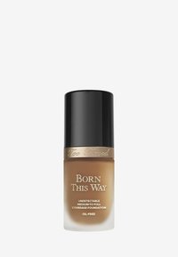 Too Faced Born This Way foundationfles, cilindervorm, heldere glazen fles met een bruine vloeistof, zwarte dop en gouden accenttekst. Vrij van olie.