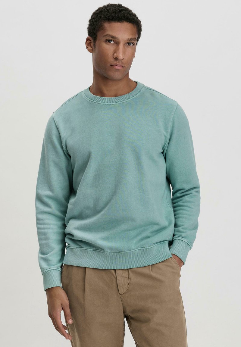 Hellblauer Rundhalsausschnitt-Sweatshirt mit gerippten Bündchen und Saum. Weiche Textur, lässige Passform. Kombiniert mit khakifarbenen Hosen, stehendes Modell.