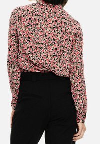 Langarmblouse van zwarte stof met een bloemenpatroon in rood, roze en crème. Achteraanzicht toont een knoopsluiting in de kraag en een aansluitend ontwerp.