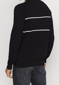 Sweat-shirt noir à col rond, avec deux bandes blanches horizontales dans le dos. Confectionné dans un tissu doux, avec des poignets et un ourlet côtelés.