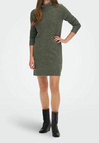 Abito lavorato a maglia verde oliva con collo alto, maniche lunghe e dettagli a coste nei polsini e nell'orlo. Indossato con stivali neri con tacco.