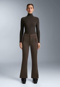 Barna, testhezálló garbó pulóver barna, bő egyenes fazonú nadrággal párosítva. A nadrágon két elülső zseb található cipzárokkal és gombos díszítéssel.