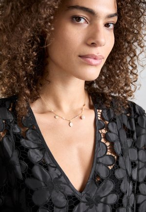 Collana d'oro con accenti di pietre rosa; top in pizzo floreale nero intricato con scollatura a V; capelli mossi naturali e trucco neutro.