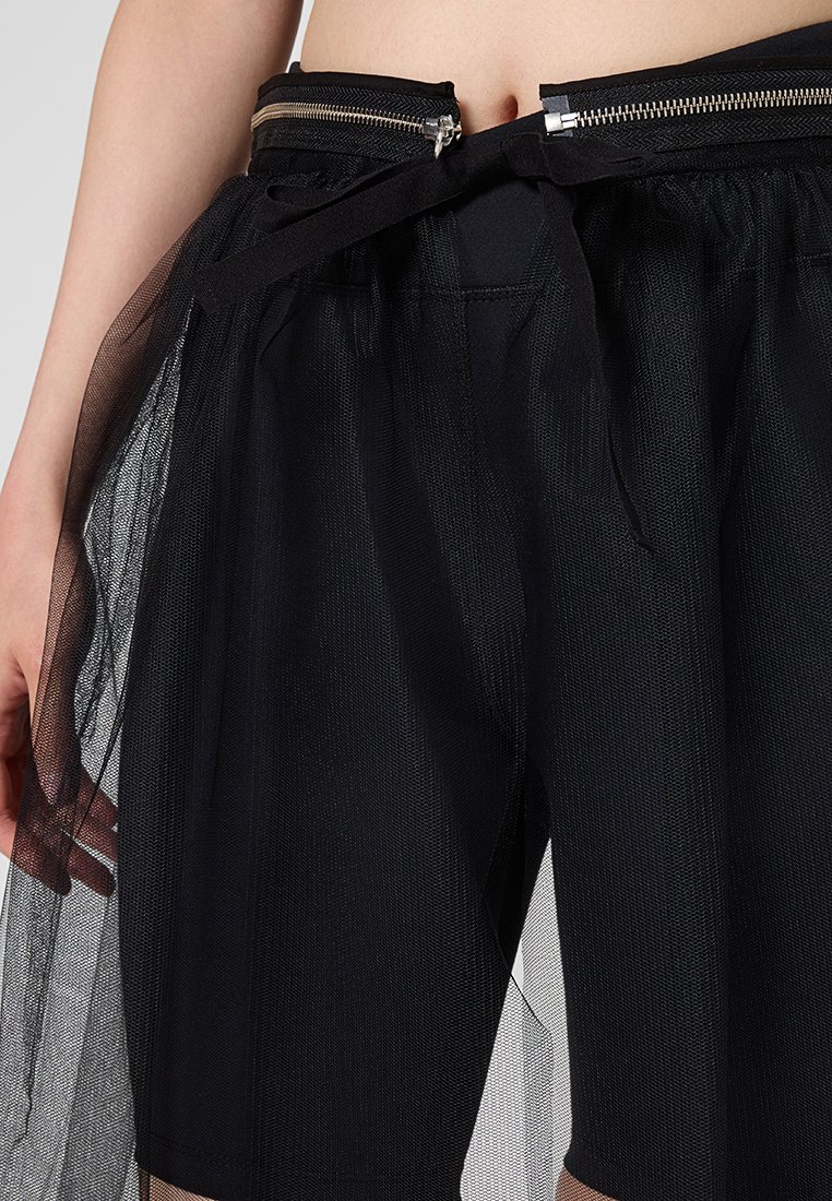 Primo piano di una gonna in rete nera sovrapposta a pantaloni corti neri con cerniere visibili e dettaglio con lacci in vita, indossata su un ventre nudo.
