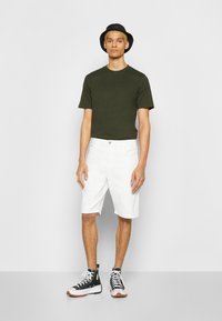 T-shirt vert olive, shorts blancs et chapeau seau noir. Baskets montantes noires et blanches avec semelles en caoutchouc. Design simple et décontracté.