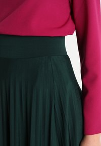 Top magenta aderente abbinato a una gonna plissettata fluida di un verde scuro. La vita è elastica, creando una transizione fluida tra i tessuti.