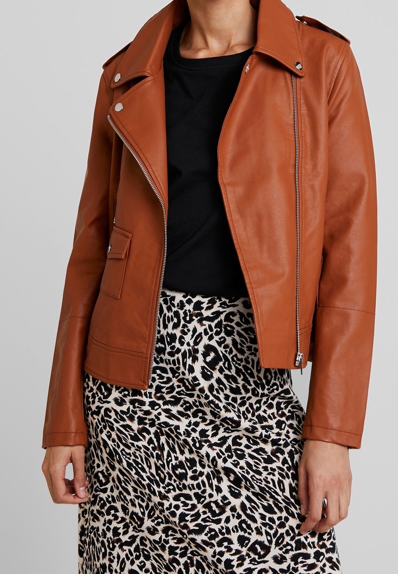Giacca da motociclista in pelle marrone con zip frontale, spalline sulle spalle e tasca laterale, indossata sopra una camicia nera e una gonna con stampa leopardata.