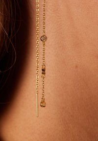 Des boucles d'oreilles pendantes en or présentent une fine chaîne avec une barre verticale géométrique et un accent rond avec une pierre transparente, sur un ton de peau.