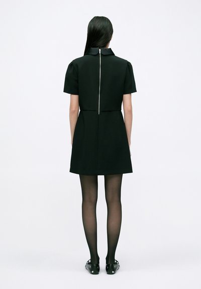 Rochie neagră cu mânecă scurtă, guler înalt și fermoar vizibil pe spate, asociată cu ciorapi negri transparenți și pantofi negri. Design simplu și structurat.