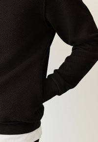 Felpa nera a coste con design aderente, scollo rotondo e maniche lunghe. Presenta uno strato di orlo bianco sul fondo.