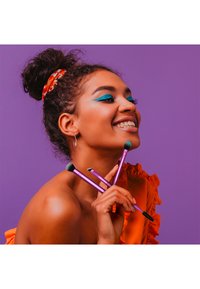 Jeune femme avec un maquillage des yeux bleu vif et un haut orange sourit en tenant trois pinceaux de maquillage violets sur un fond violet.