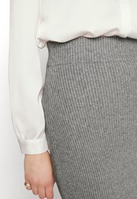 Grå ribbad stickad kjol med ett brett elastiskt midjeband, ihopparad med en vit blus med långa ärmar. Synlig hand som bär en guldring.