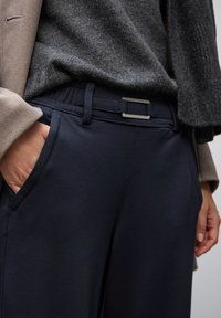 Detalle de una persona vistiendo pantalones marinos con un cinturón de hebilla rectangular de plata, un suéter gris texturizado y una manga de abrigo beige.