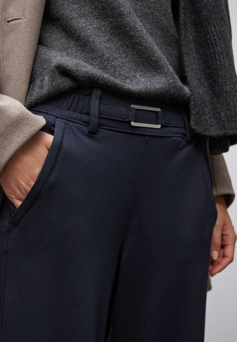 Detalle de una persona vistiendo pantalones marinos con un cinturón de hebilla rectangular de plata, un suéter gris texturizado y una manga de abrigo beige.