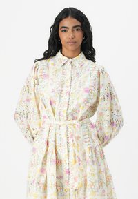 Lätt blommig bomullsklänning med knappframsida, krage och midja med bälte. Har puffärmar och intrikata broderidetaljer.