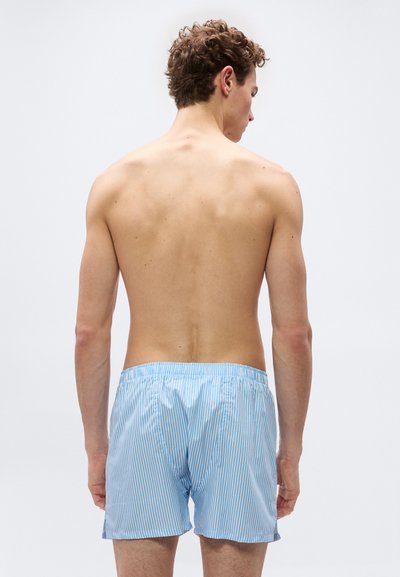 Νέος άνδρας με σγουρά μαλλιά, γυρισμένος από την πλάτη, χωρίς πουκάμισο, φοράει ανοιχτό μπλε και λευκά ριγέ μπόξερ πάνω σε απλό ανοιχτό φόντο.