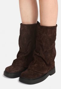 Stivali in suede marrone di altezza media, con suole nere spesse, indossati su gambe nude, mostrano una vestibilità morbida e rilassata intorno alle caviglie.