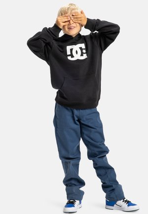 Zwarte hoodie met DC-logo, voorzak, blauwe baggy broek en witte sneakers met blauwe accenten. Kind in een speelse pose waardoor het gezicht niet zichtbaar is.