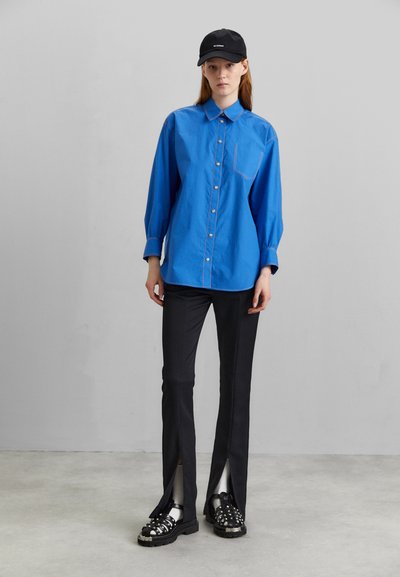 Μπλε πουκάμισο με μακριά μανίκια, κουμπιά και τσέπη στο στήθος, συνδυασμένο με μαύρες tailored παντελόνες με πλευρικές σχισμές και μαύρα σανδάλια με χοντρή σόλα.