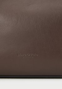 Sac en cuir marron avec une texture lisse, arborant un logo en relief « Marc O'Polo » et une date d'établissement. Design simple et minimaliste.