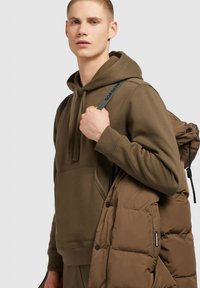 Oli Green hoodie met kangoeroezak, verstelbare trekkoorden, gedragen met een bruine pufferjack. Gladde stof, losse pasvorm, minimale details.