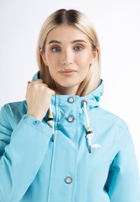 Jachetă impermeabilă de culoare albastru deschis, cu guler înalt, șnururi de ajustare, nasturi cu capsenă și detalii cu logo. Textură netedă și croială lejeră.